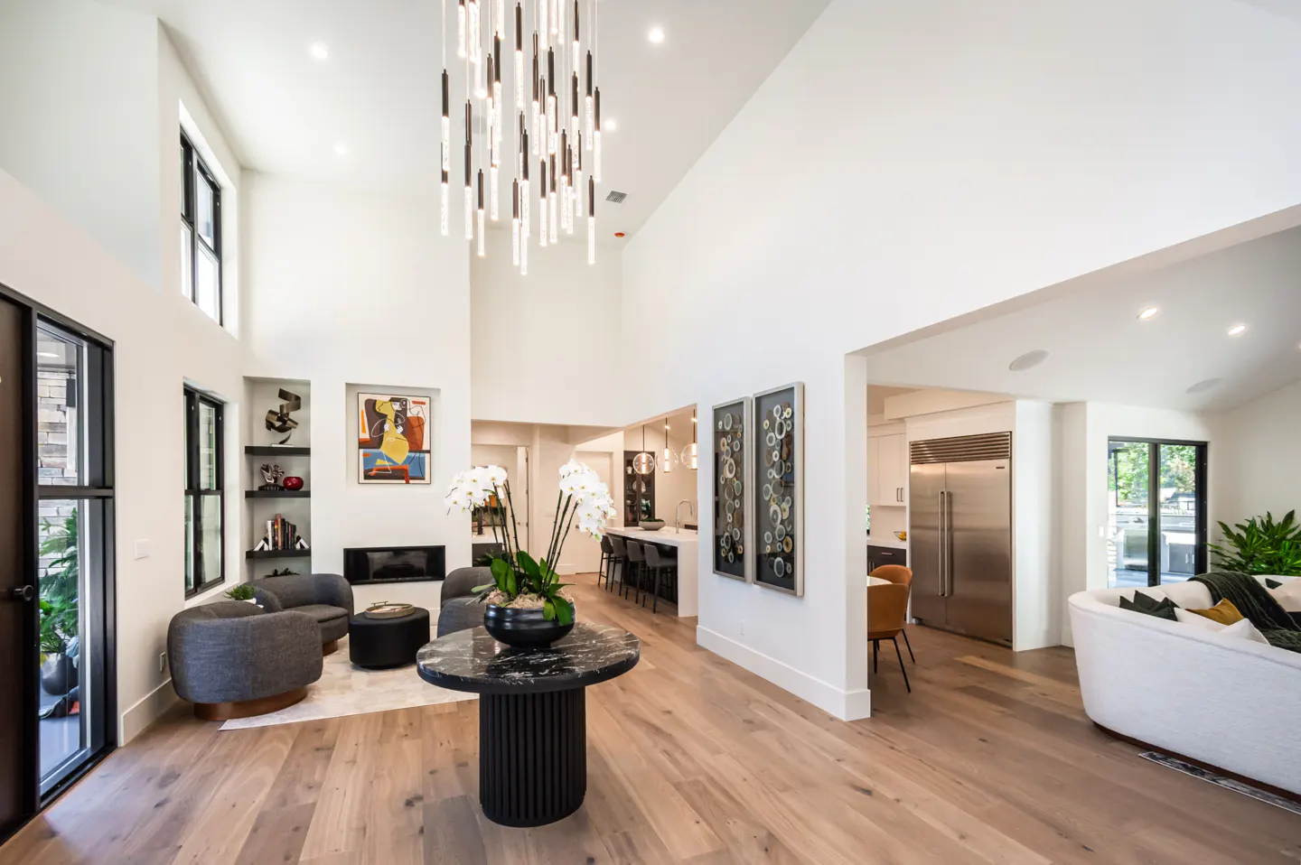 Bright, open-concept living space with hardwood floors, white walls, and black-framed windows. A modern chandelier hangs above a black marble table with orchids.