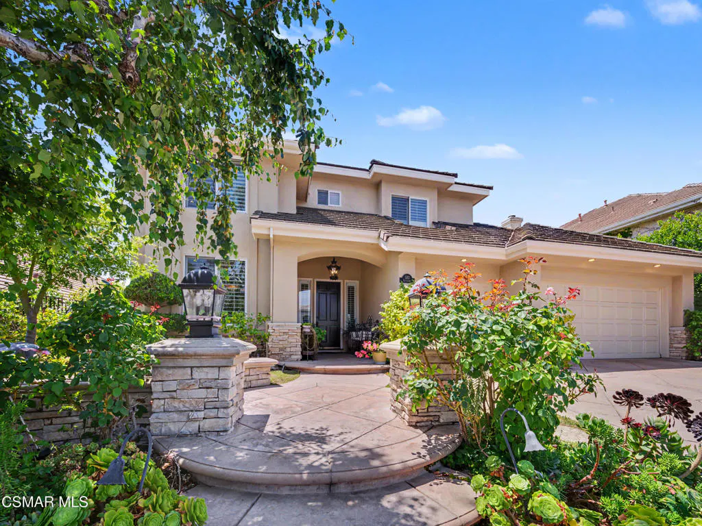 Two-story beige suburban home with a stone-accented entryway, curved walkway, lush landscaping, and a two-car garage.