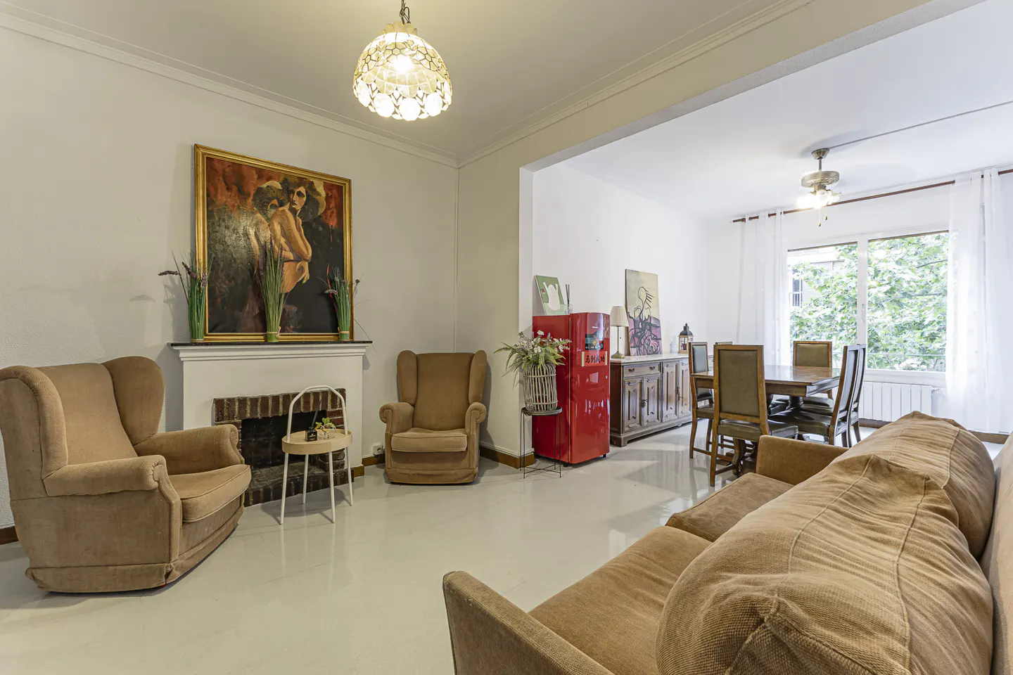 A living room with tan armchairs, a brown sofa, and a red refrigerator. A dining room is visible in the background.