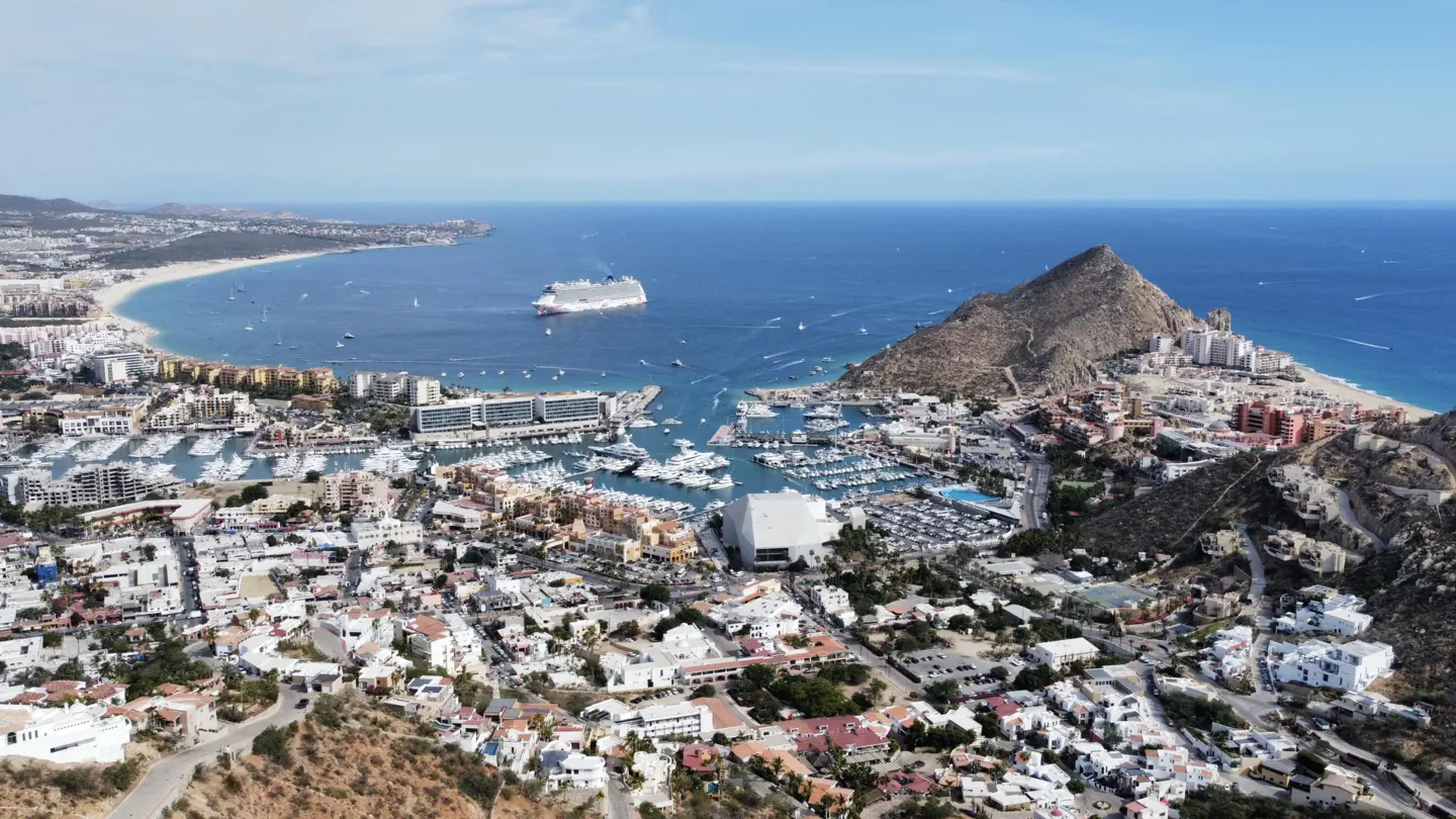 Aerial view of Cabo San Lucas, Mexico, with a harbor full of boats, a cruise ship in the bay, and a mountain backdrop.