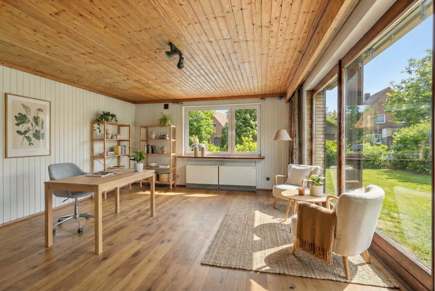 Bright sunroom with wood ceiling, large windows, and hardwood floors. A desk, chairs, and shelves furnish the space. Outside, green trees and a house are visible.