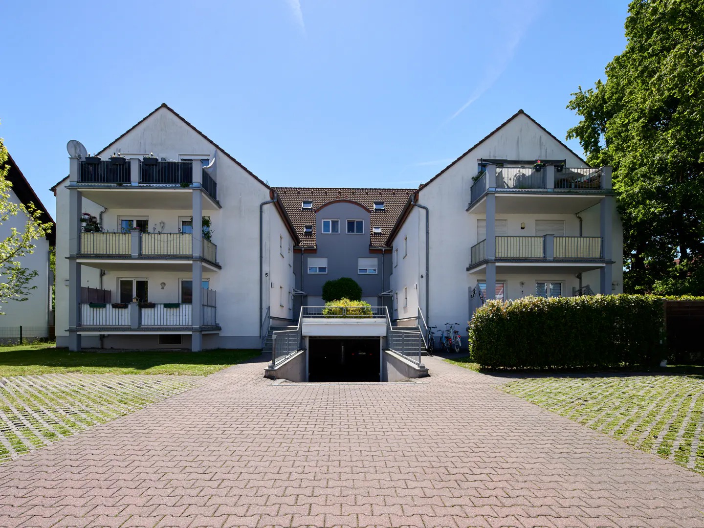 Exterior view of a white apartment building with balconies and a brick driveway on a sunny day.