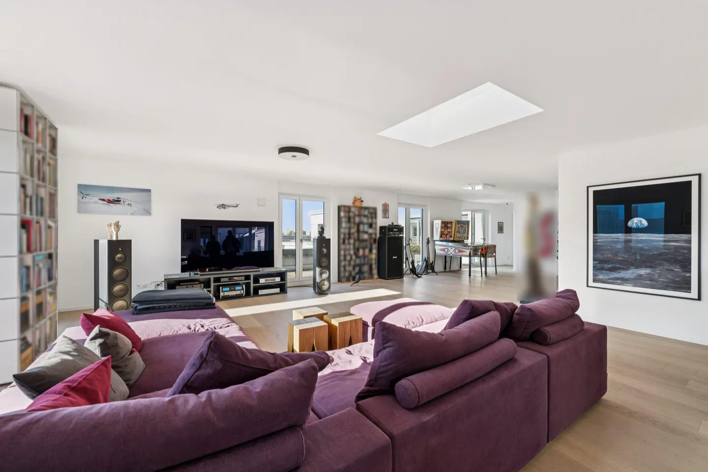 Bright, open living room with a large purple sectional sofa, TV, speakers, and a bookcase. A pinball machine is visible in the background.