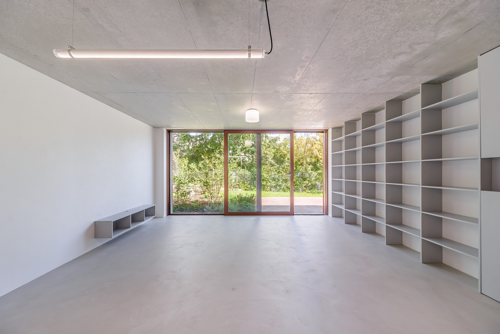 Minimalist room with concrete ceiling, white walls, large window with green view, and built-in gray shelving.