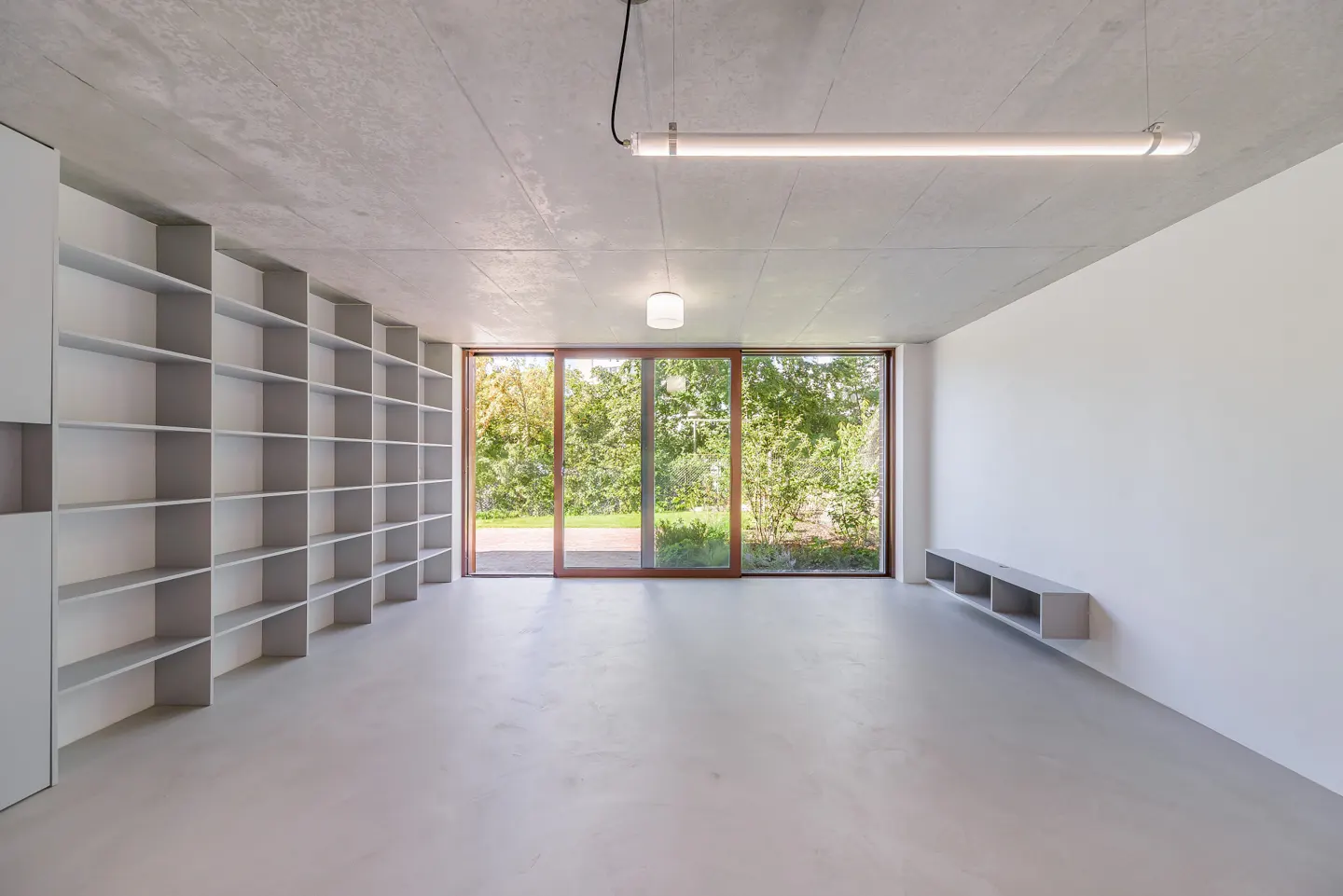 Minimalist room with concrete ceiling, white walls, large window with green view, and built-in gray shelving.