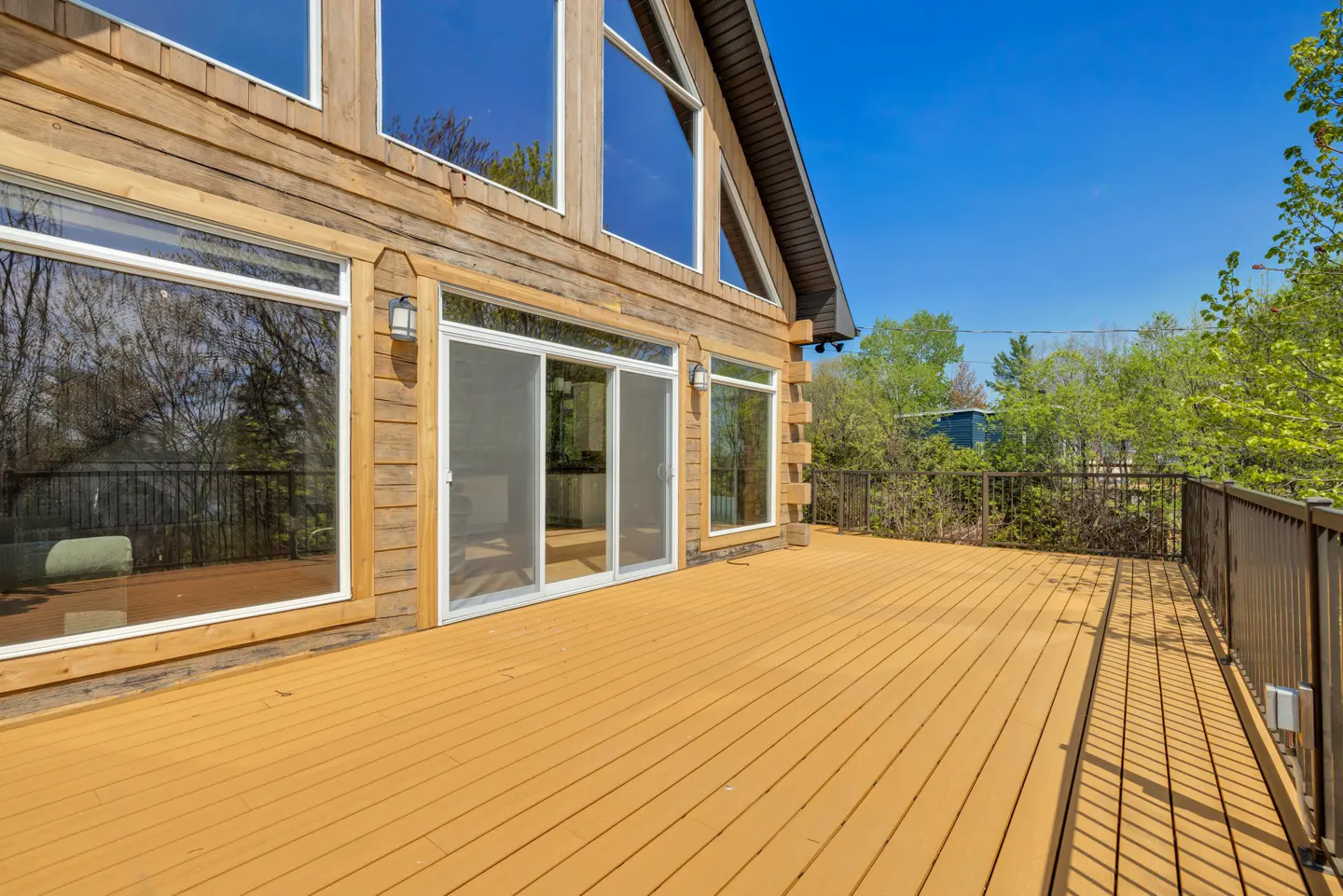 Exterior view of a log cabin with a large wooden deck, sliding glass doors, and large windows under a clear blue sky.