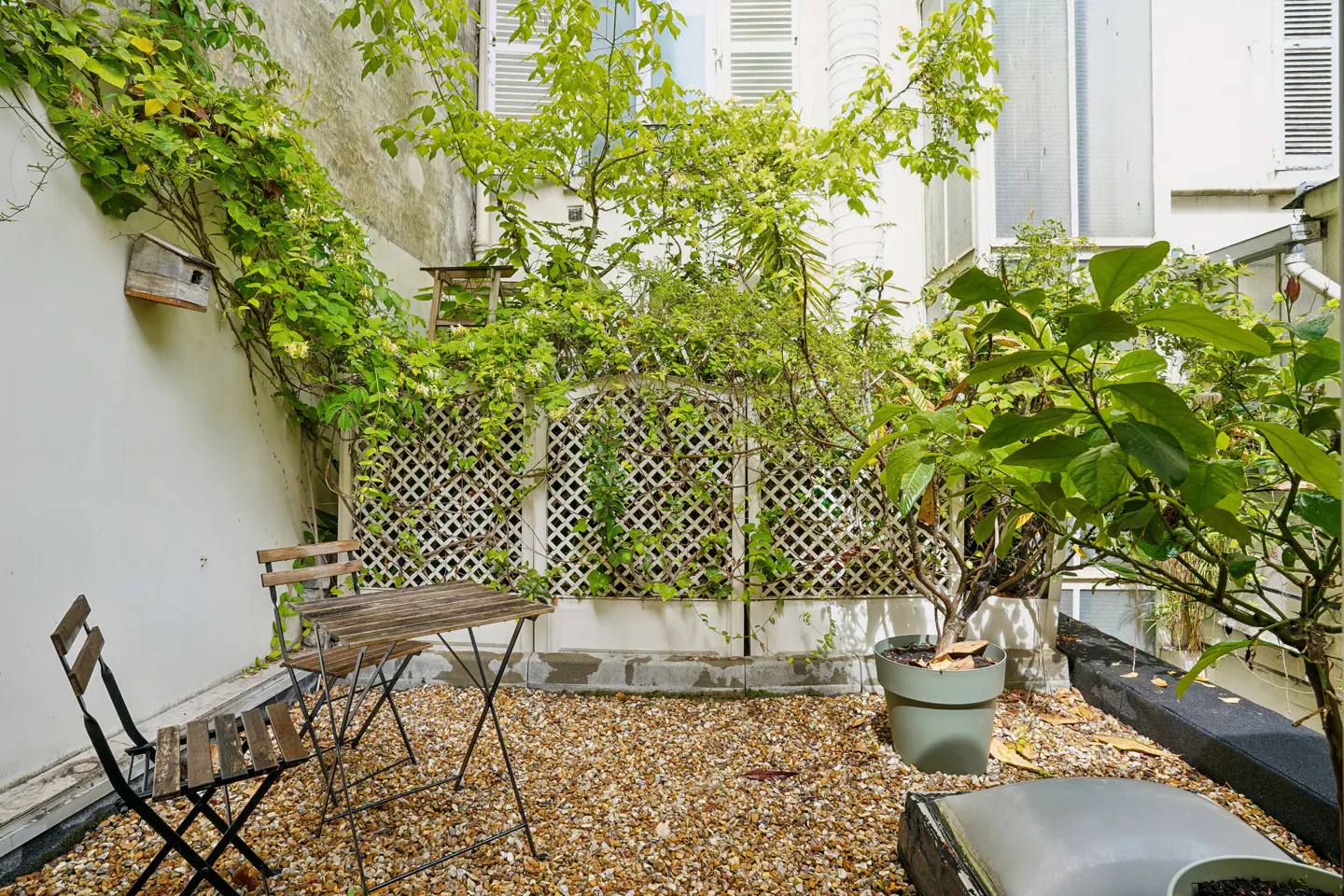 A small outdoor patio with a wooden table and chairs, gravel ground, and climbing plants on the walls.