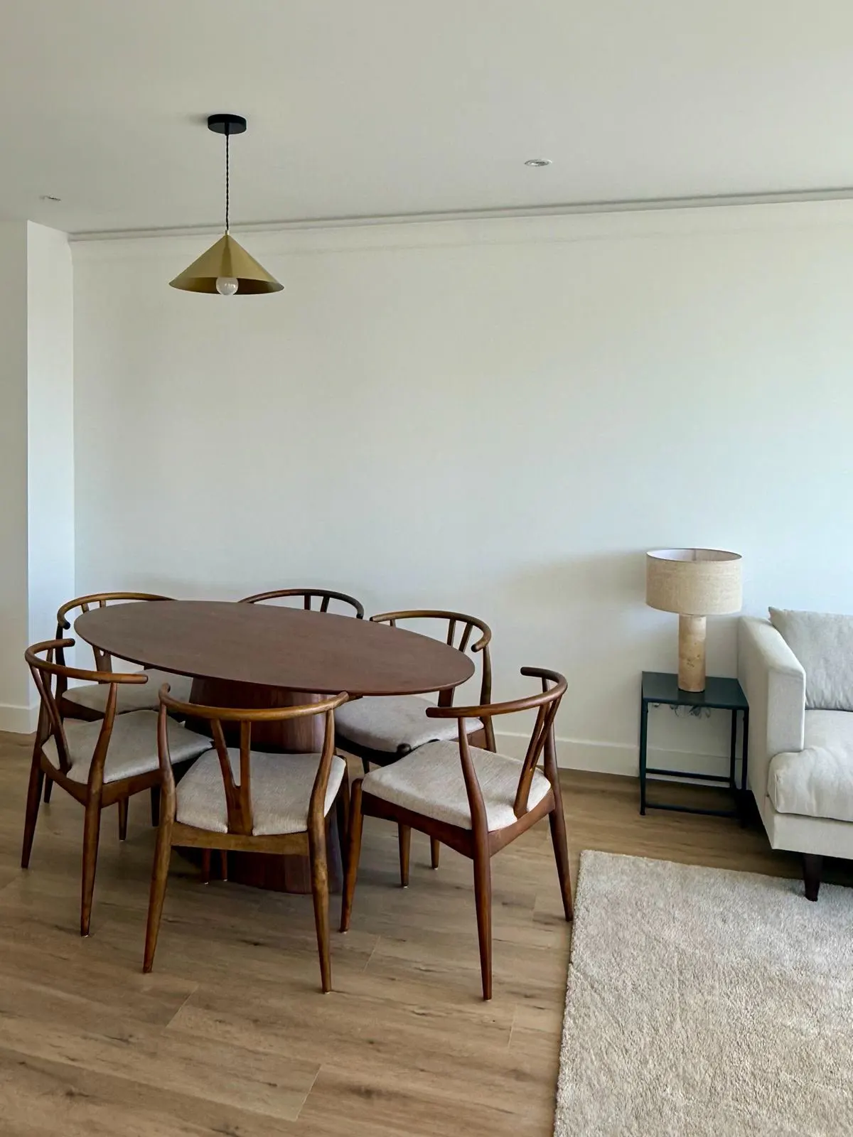 Modern dining room with an oval wood table, four wooden wishbone chairs, a brass pendant light, and a beige sofa nearby.