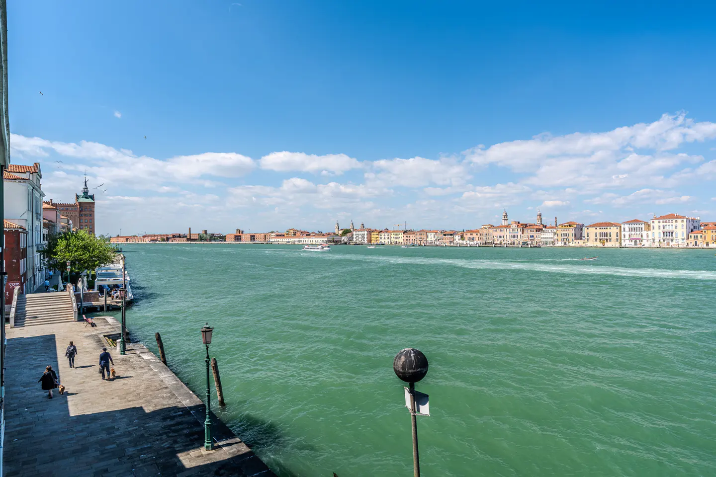 Venice cityscape on a sunny day. Turquoise water, buildings in the distance, and people walking along the stone walkway.
