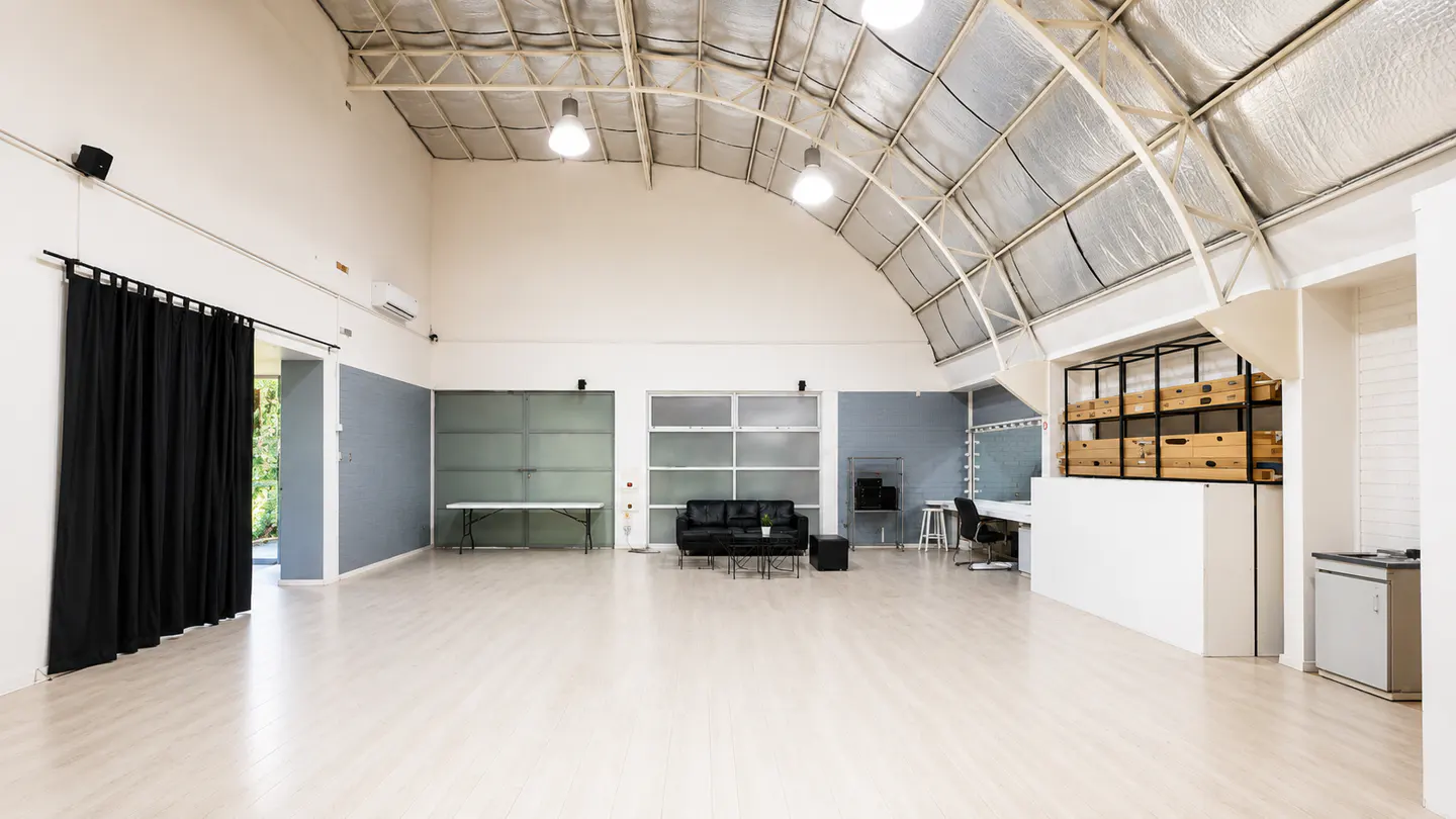 Bright, empty studio with a curved metal roof, white walls, and light wood floors. Black curtains, a sofa, and a makeup station are visible.