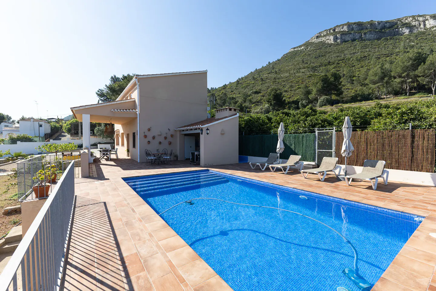 A tan house with a blue tiled pool and lounge chairs sits in front of a green, tree-covered mountain.