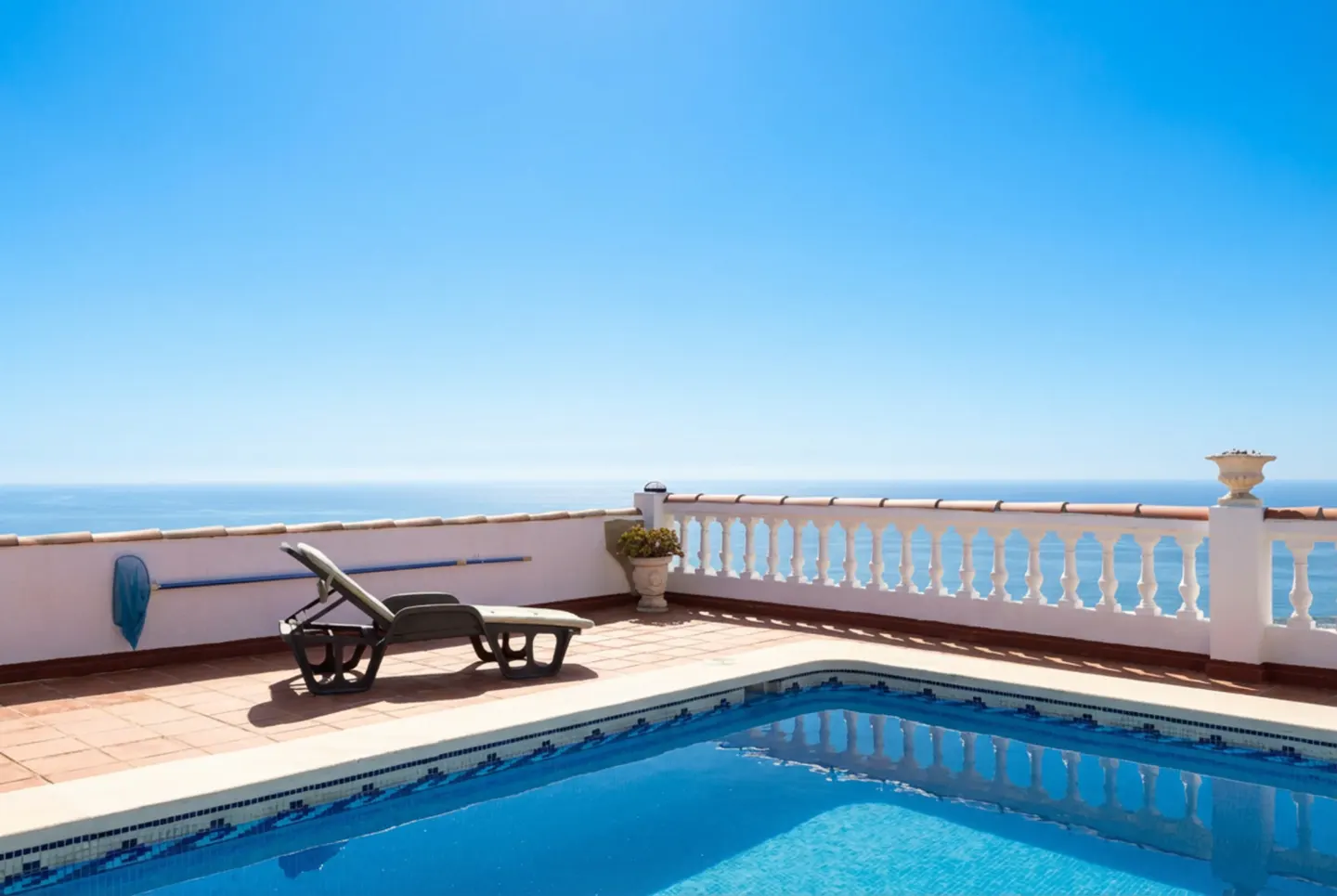 A blue tiled pool with a lounge chair and ocean view under a clear blue sky.