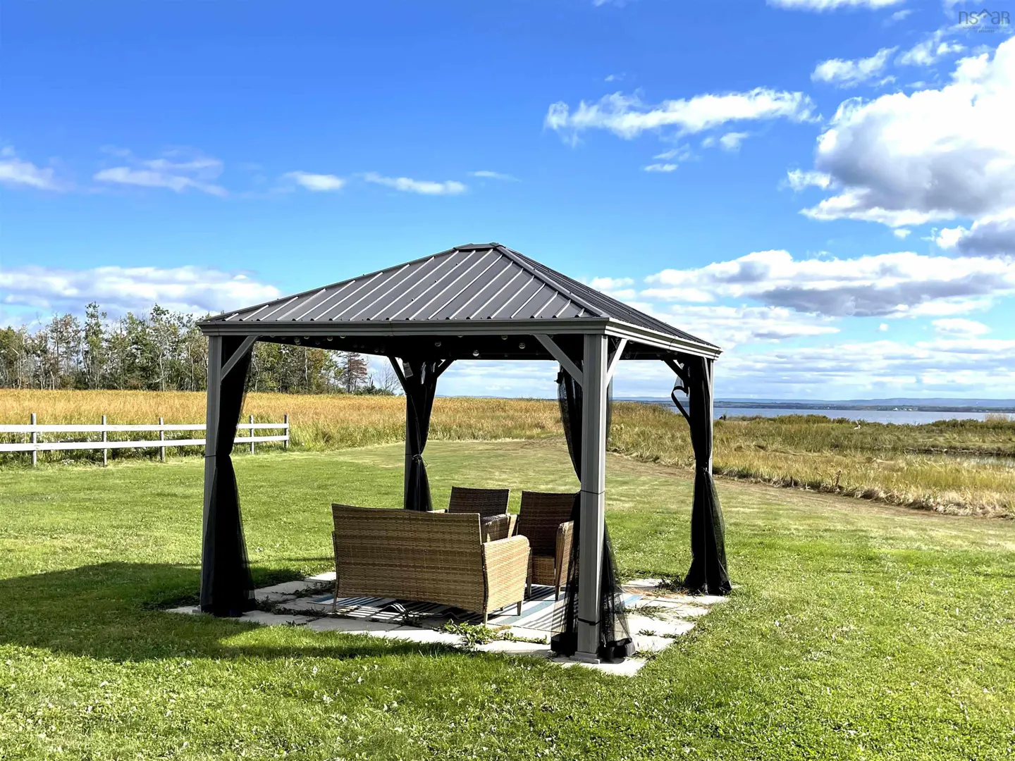 Outdoor gazebo with wicker furniture on a green lawn, under a blue sky with clouds. A field and water are in the background.
