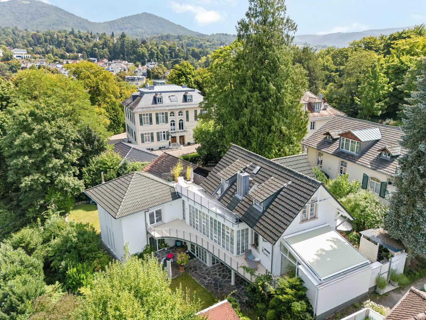 Aerial view of a white house with a gray roof, surrounded by green trees and mountains in the background.
