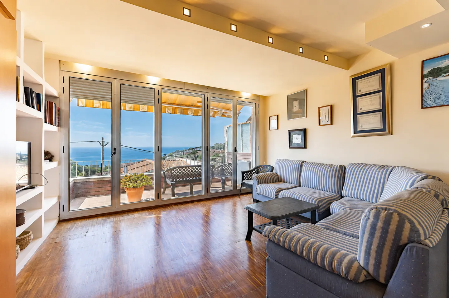 Living room with blue and white striped sectional sofa, wooden floors, and large glass doors opening to a sea-view balcony.