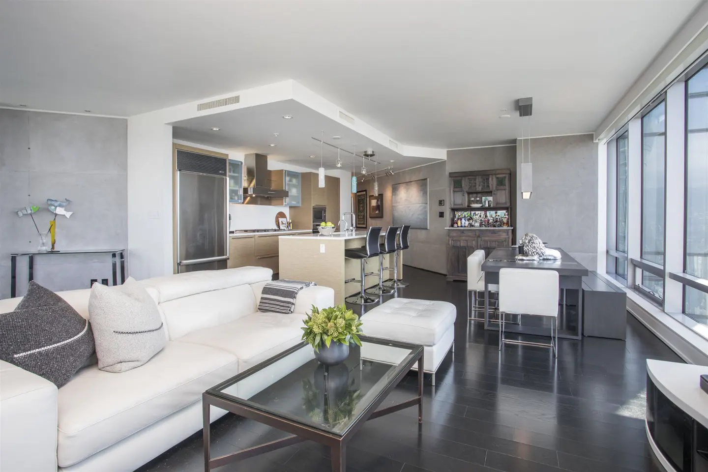 Open-concept condo interior with a white sofa, kitchen island, dining table, and floor-to-ceiling windows.