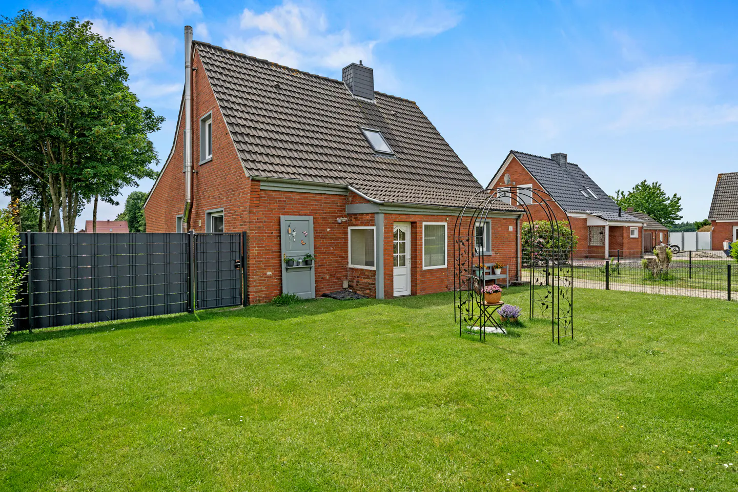 Red brick house with a dark tiled roof, a green lawn, a metal garden arch, and a black privacy fence under a blue sky.