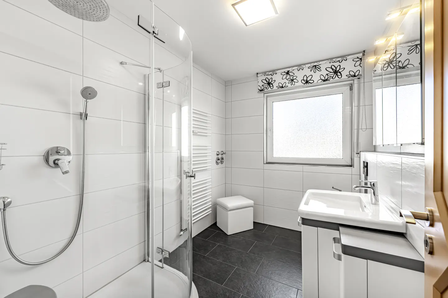 Bright bathroom with white tile walls, dark floor, glass shower, sink vanity, and window with floral valance.