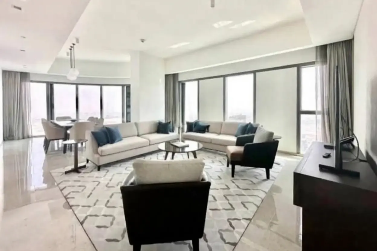 Bright, modern living room with beige sofas, blue pillows, and large windows with gray curtains. A dining table is visible in the background.