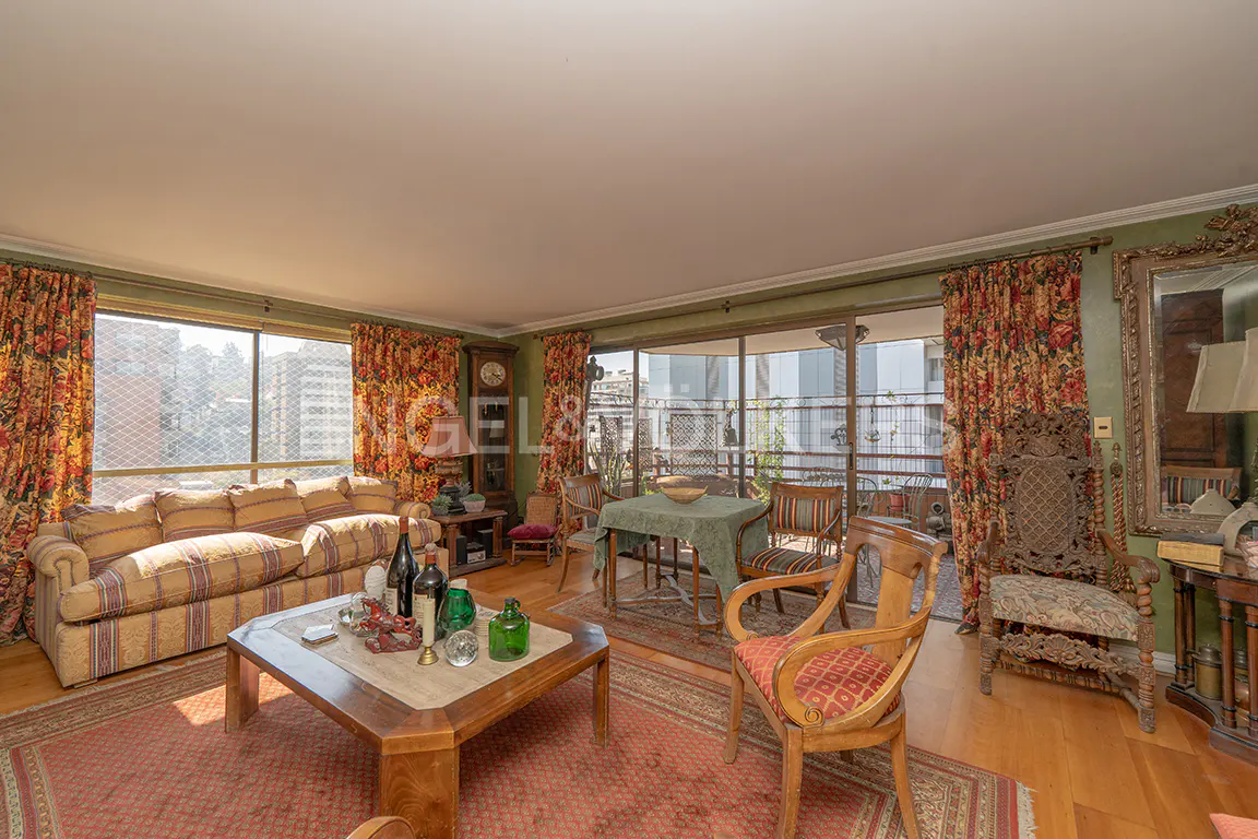 A living room with a sofa, table, chairs, and floral curtains. A balcony is visible through the sliding glass doors.