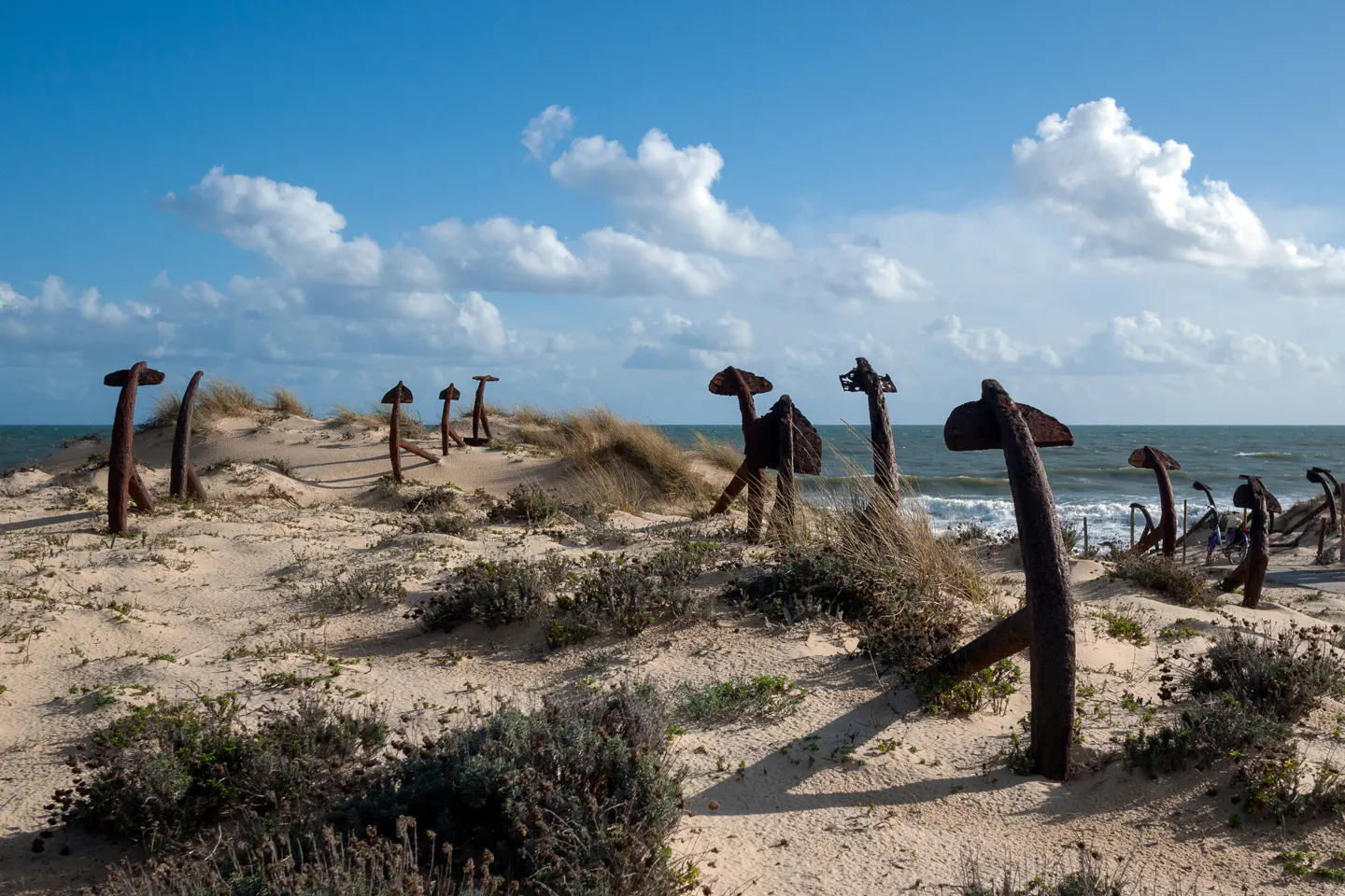 Sandy beach with rusty anchors standing upright, facing the ocean under a blue sky with scattered clouds.