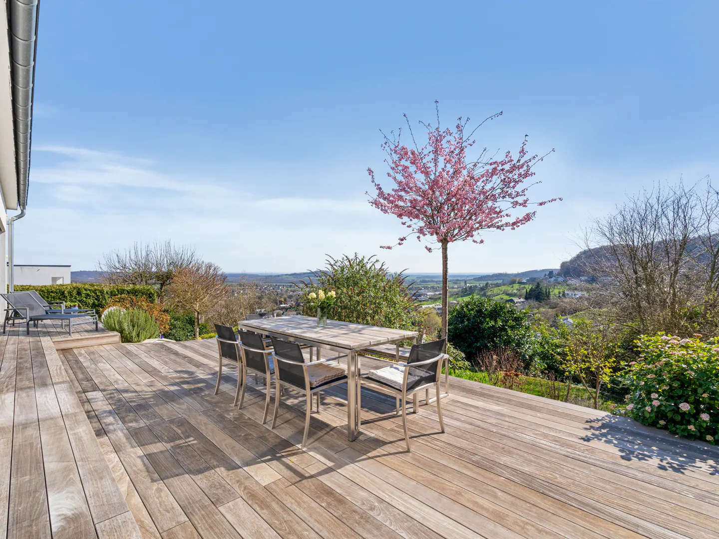 Outdoor wooden deck with a table and chairs overlooking a scenic landscape with a pink blossoming tree.