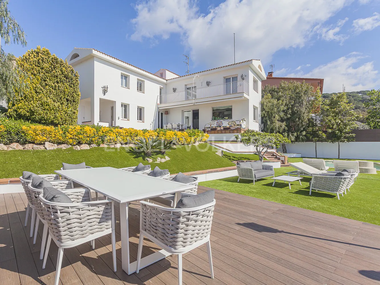 Backyard view of a two-story white house with a patio set on a wooden deck and lawn furniture on the grass.