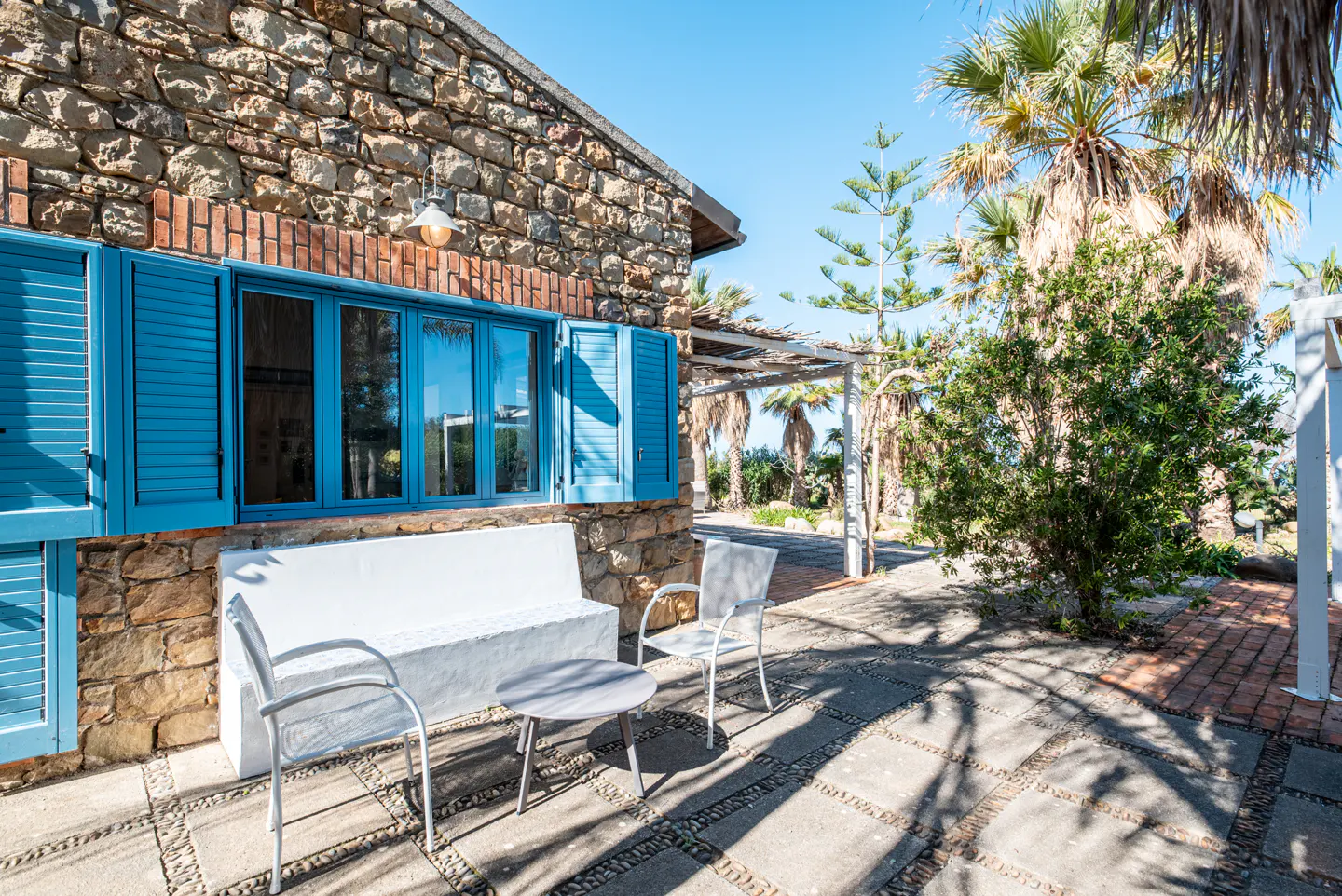 Stone house with blue shutters, white bench, and chairs on a patio. Palm trees and greenery in the background under a clear blue sky.