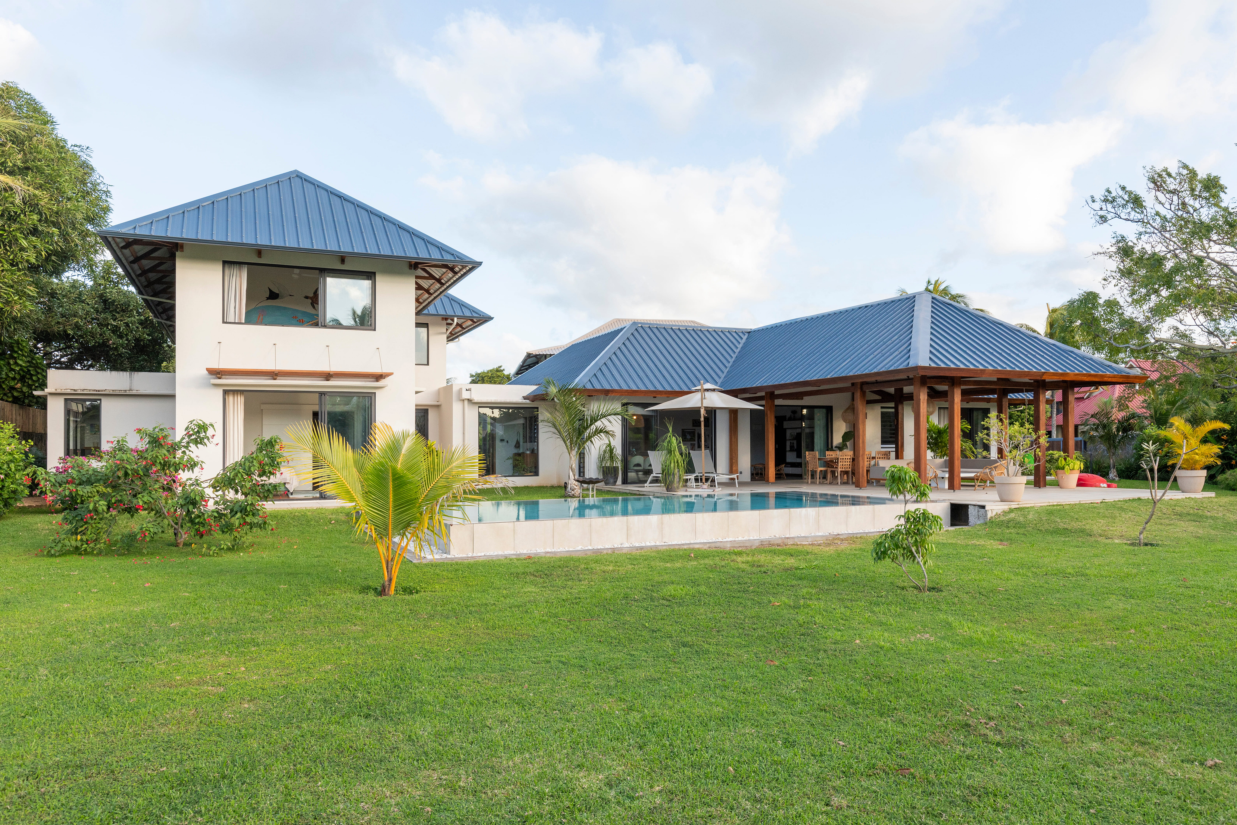 Two-story white villa with a blue metal roof, swimming pool, and lush green lawn under a bright, partly cloudy sky. [AI-generated text]