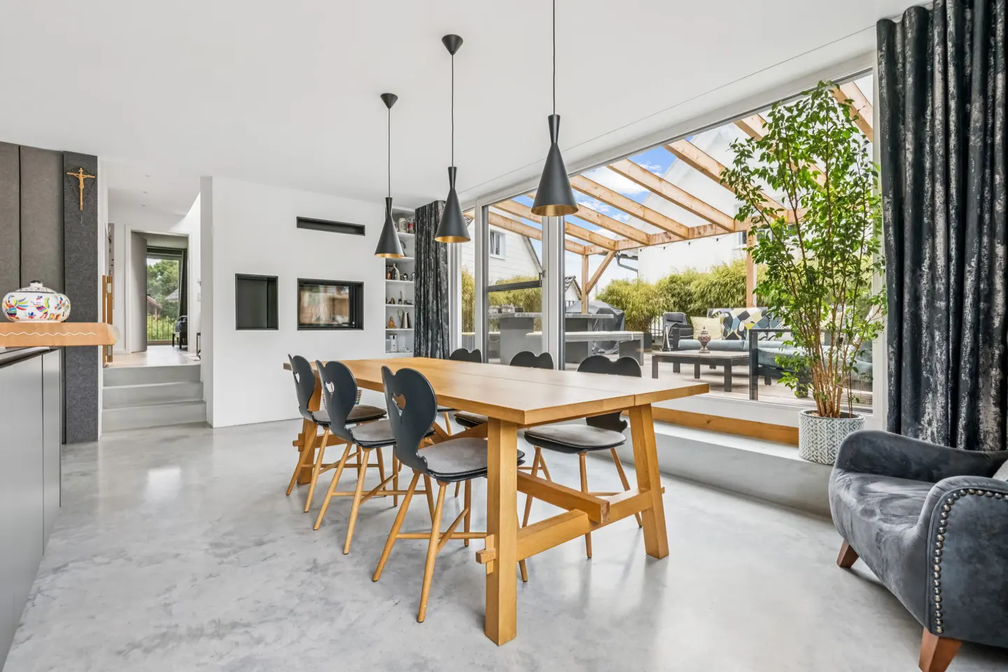 Modern dining room with wooden table, black chairs, concrete floors, and large windows overlooking a patio with a pergola.