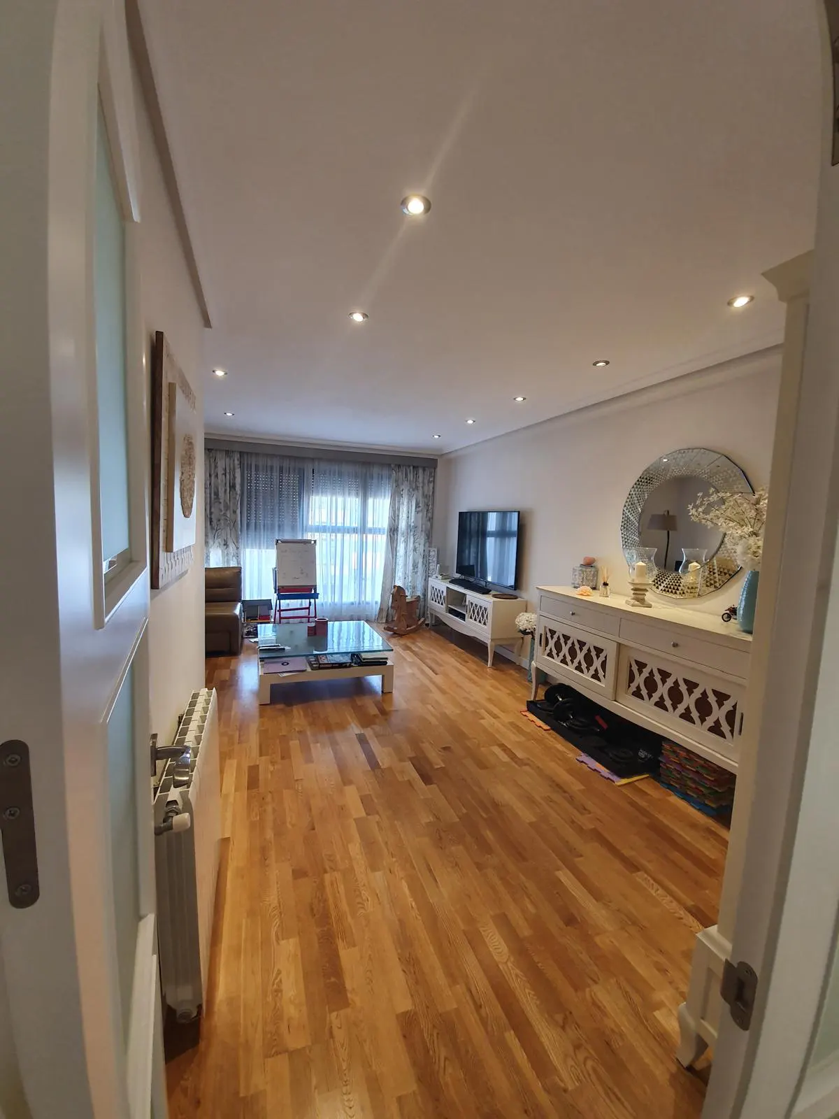 A bright living room with hardwood floors, white walls, and recessed lighting. A large window with patterned curtains is visible.