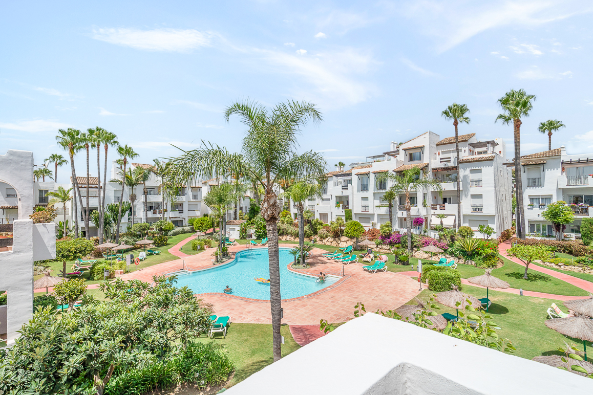 High-angle view of a resort pool surrounded by white apartment buildings, palm trees, green lawns, and lounge chairs. [AI-generated text]