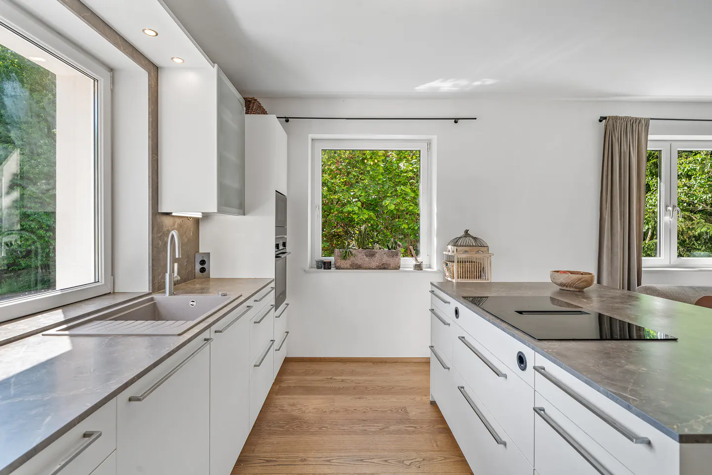 Bright kitchen with white cabinets, gray countertops, and wood floors. Windows overlook lush greenery. Island with cooktop and decorative birdcage.
