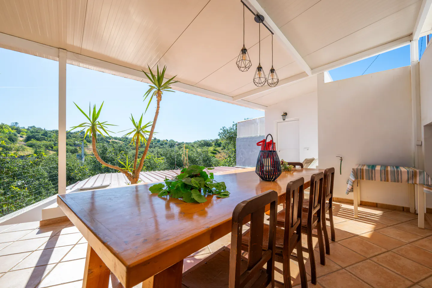 Outdoor dining area with a long wooden table, chairs, and a view of green hills under a bright blue sky.