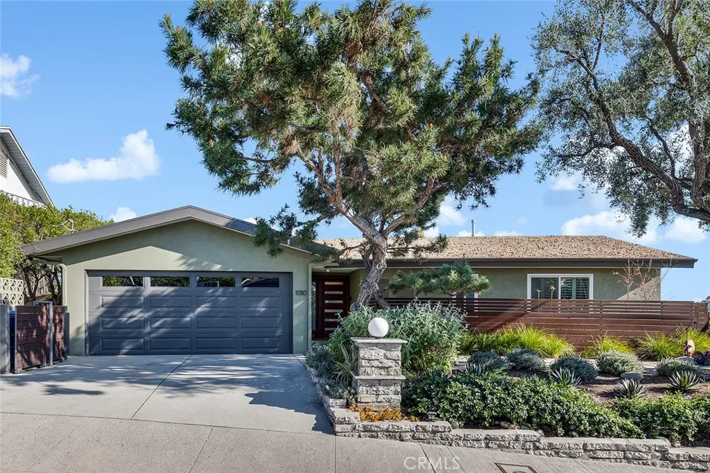 A single-story home with a gray garage door, olive green exterior, and a brown roof under a blue sky. A large tree is in the front yard.