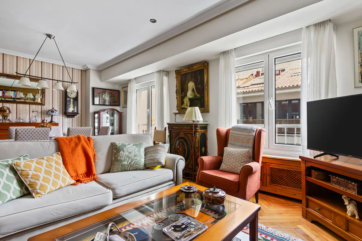 Cozy living room with a beige sofa, orange throw, patterned pillows, an armchair, and a glass-top coffee table.