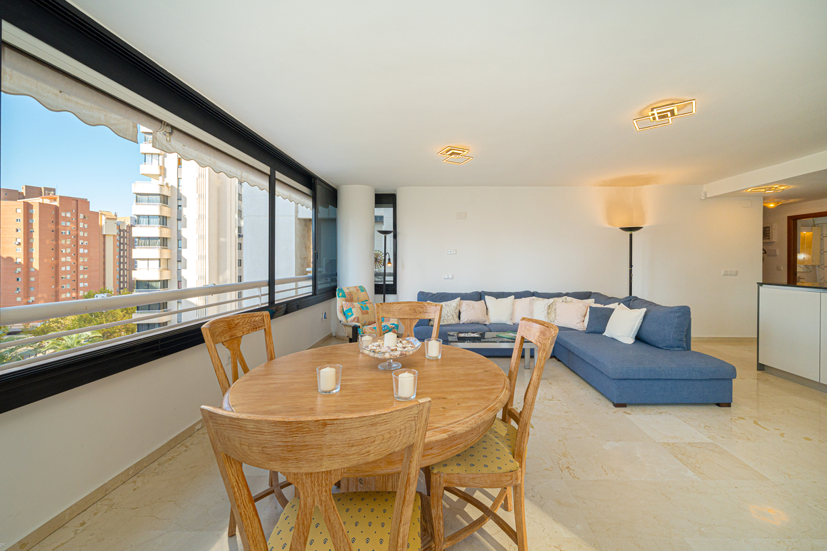 Bright living room with a round wooden dining table, blue sectional sofa, and large windows overlooking city buildings. [AI-generated text]