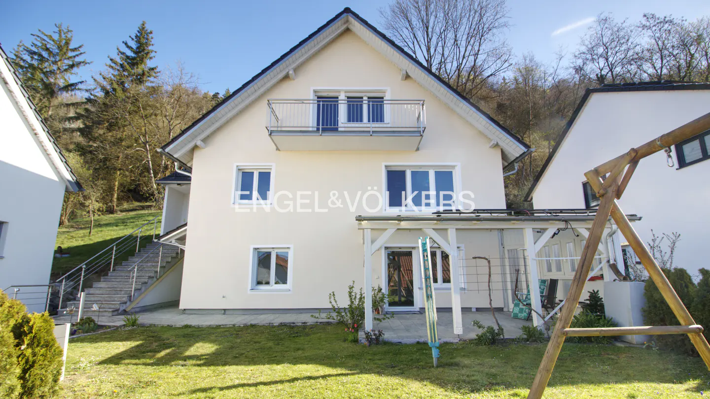 Two-story house with a balcony, beige exterior, white trim, and a green lawn. A wooden swing set is in the yard. Trees in the background.