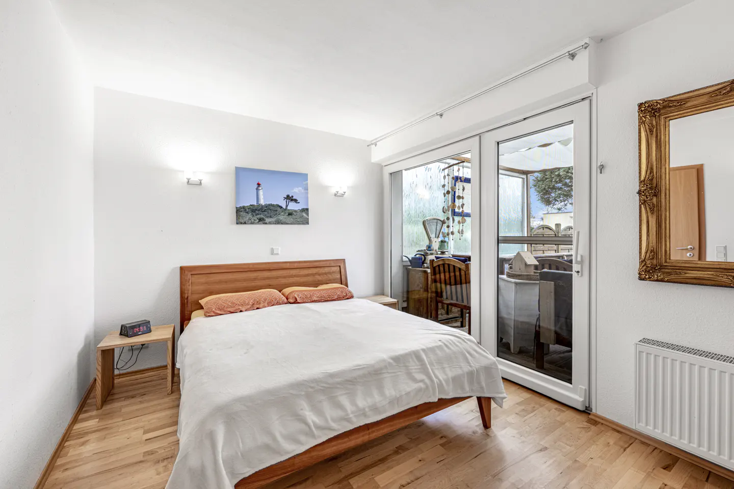 Bright bedroom with wood floors, white walls, and a double bed. Sliding glass doors lead to a sunroom. A gold-framed mirror hangs on the wall.