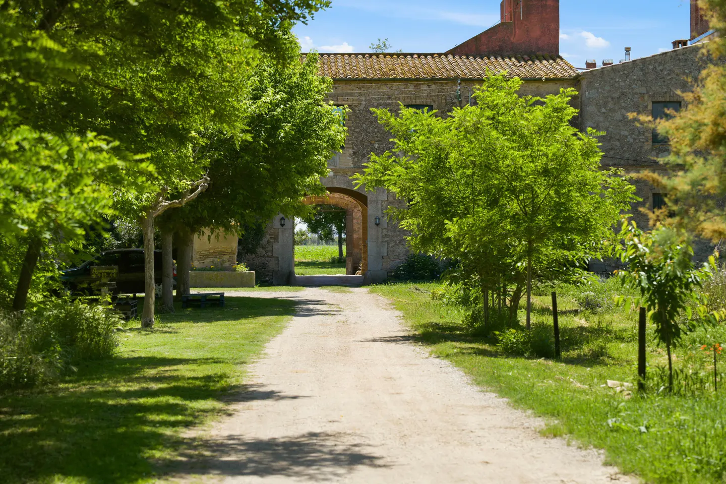 Gravel driveway leads through stone archway into a green field. Lush trees frame the path to the building.