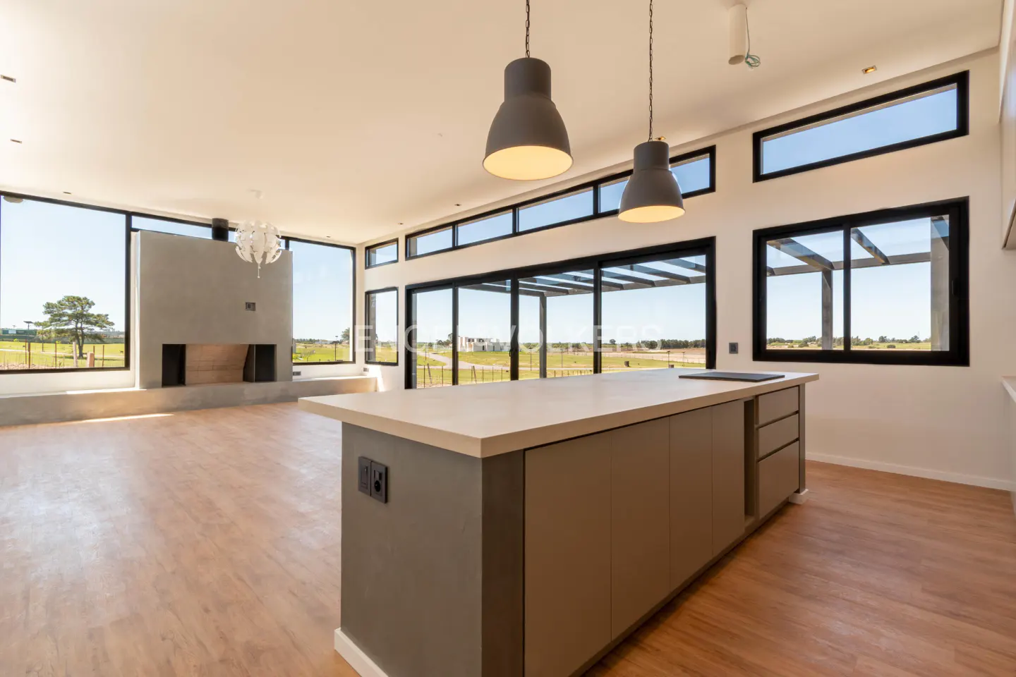 Bright, modern kitchen with a gray island, wood floors, and large windows overlooking a green field. Two gray pendant lights hang above the island.