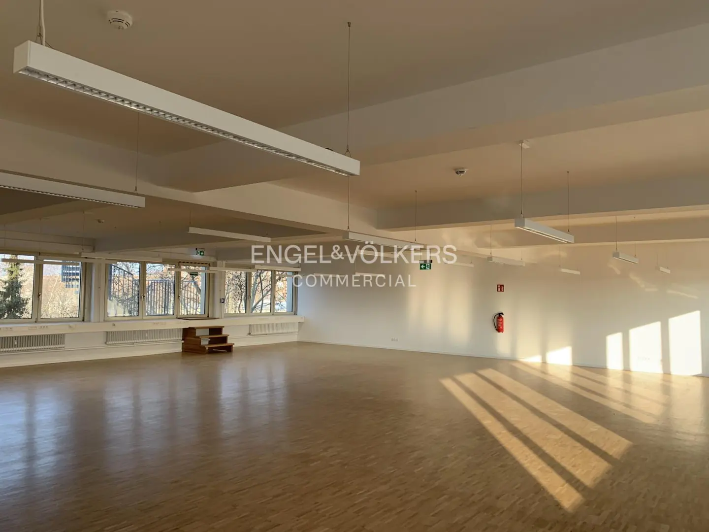 Bright, empty commercial space with wood floors, white walls, and large windows. Overhead lighting and Engel & Völkers logo visible.