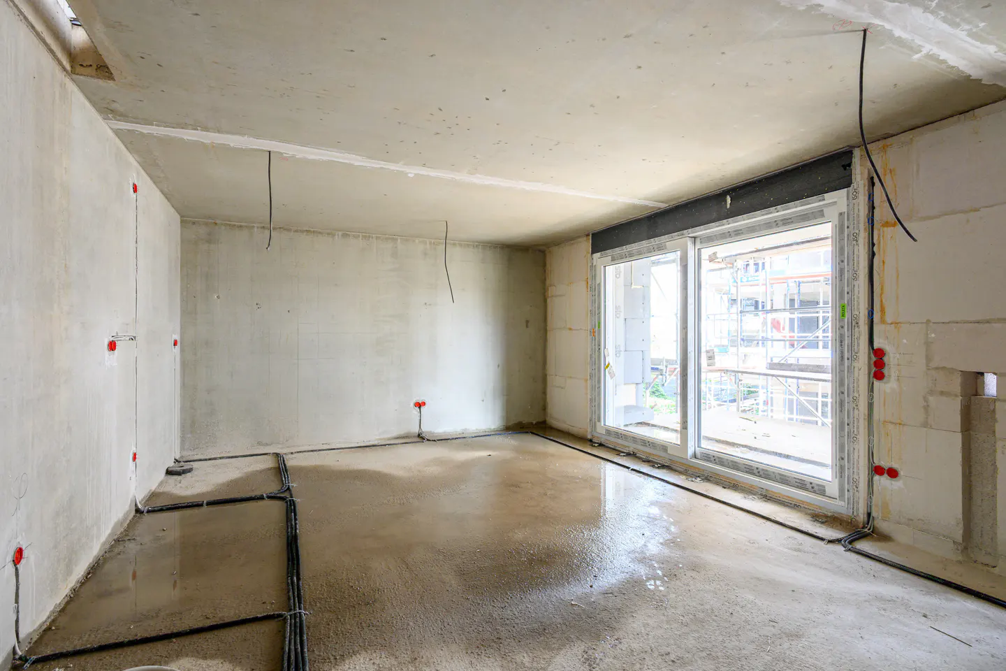 Unfinished room with concrete walls, floor, and ceiling. Black cables run along the floor and walls. A large window shows a building under construction.