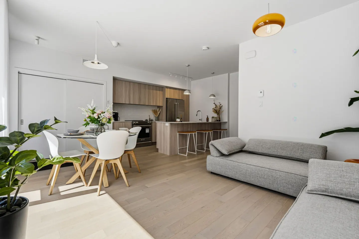 Bright, modern apartment with a gray sofa, dining table with white chairs, and a kitchen island with stools. Light wood floors and white walls.
