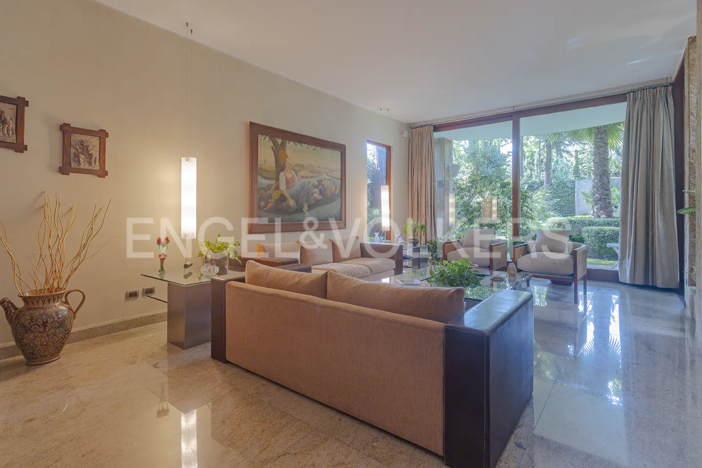 Living room with tan sofas, marble floors, and large windows overlooking a lush green garden. Artwork adorns the walls.