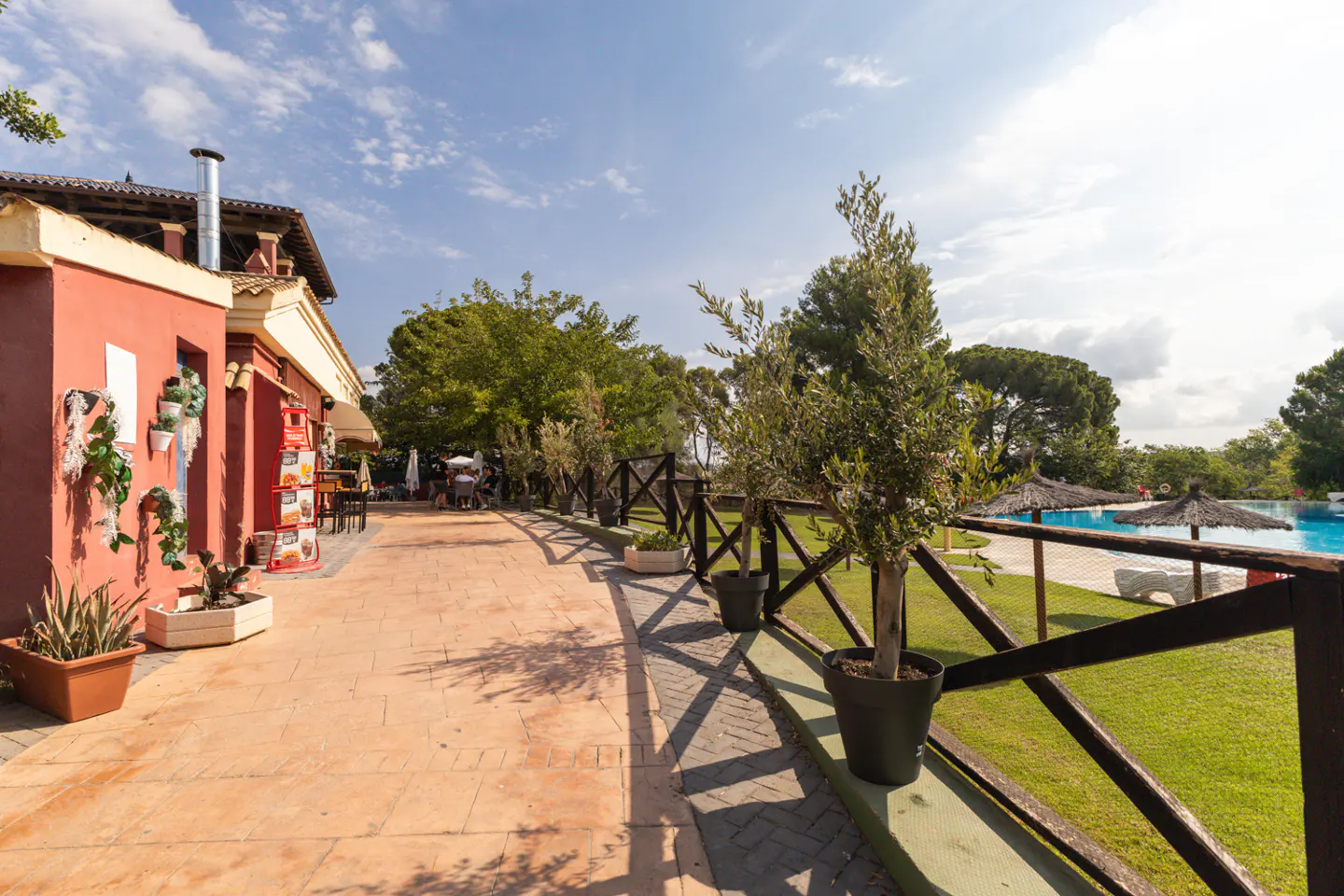 Outdoor patio with terracotta tiles, a red building, and a wooden fence overlooking a pool and green lawn.