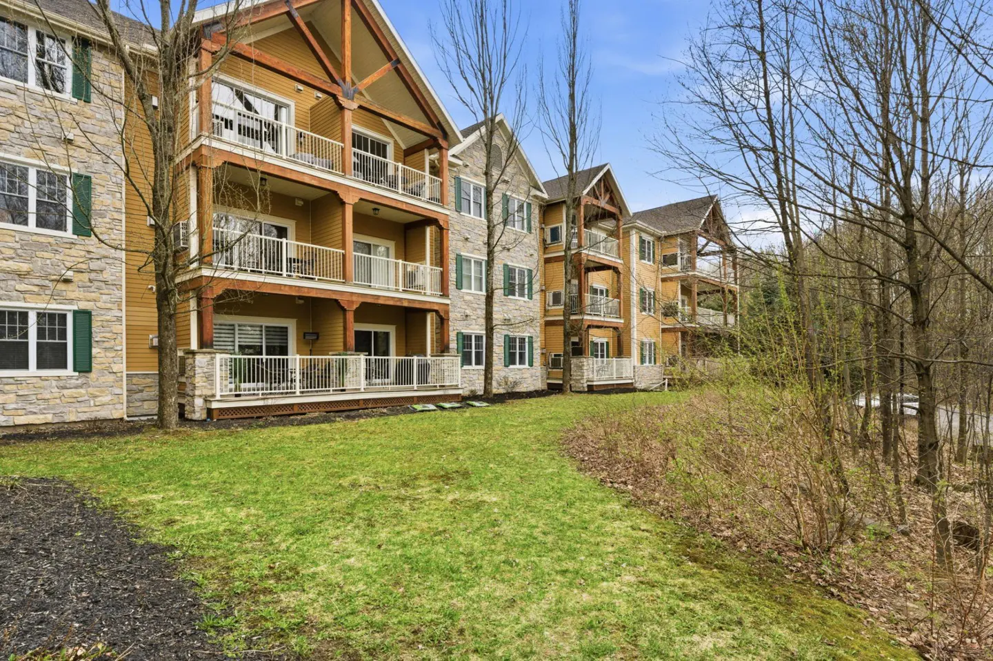 Three-story condos with balconies, stone and yellow siding, green shutters, and a grassy lawn.