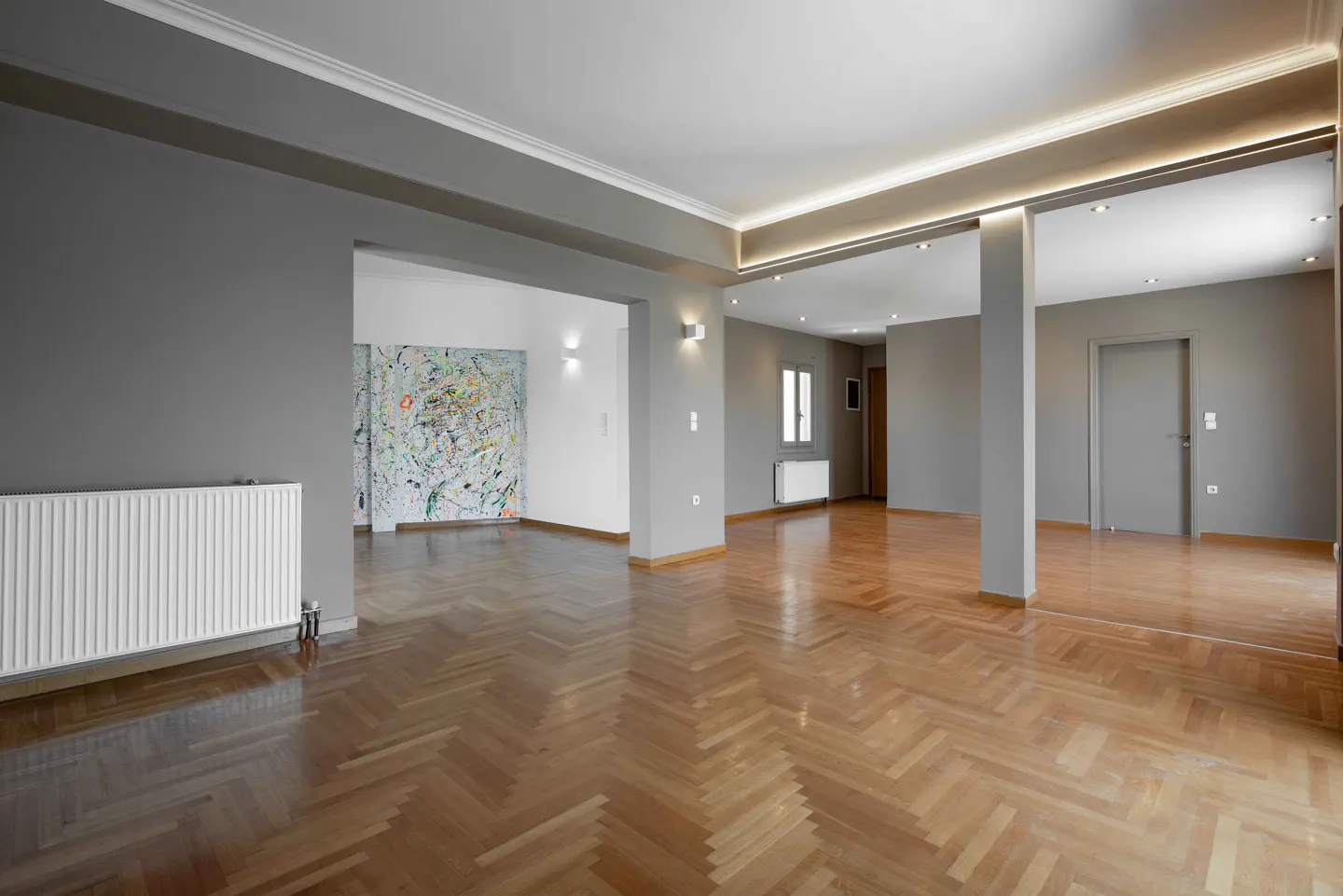 Empty room with herringbone wood floors, gray walls, and white trim. A large abstract painting is visible in the next room.