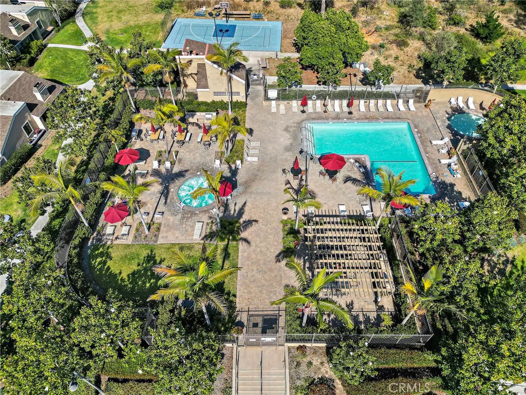 Aerial view of a community pool area with palm trees, red umbrellas, lounge chairs, and a basketball court in the background.