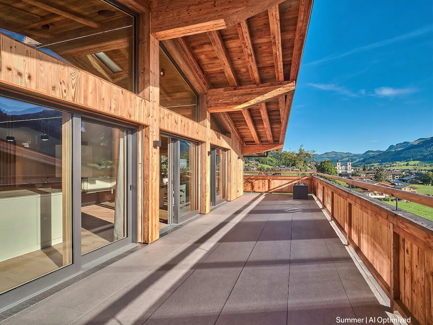 Wide balcony with wood railing and gray tile floor. Large glass doors lead inside. Distant view of town, mountains, and blue sky.